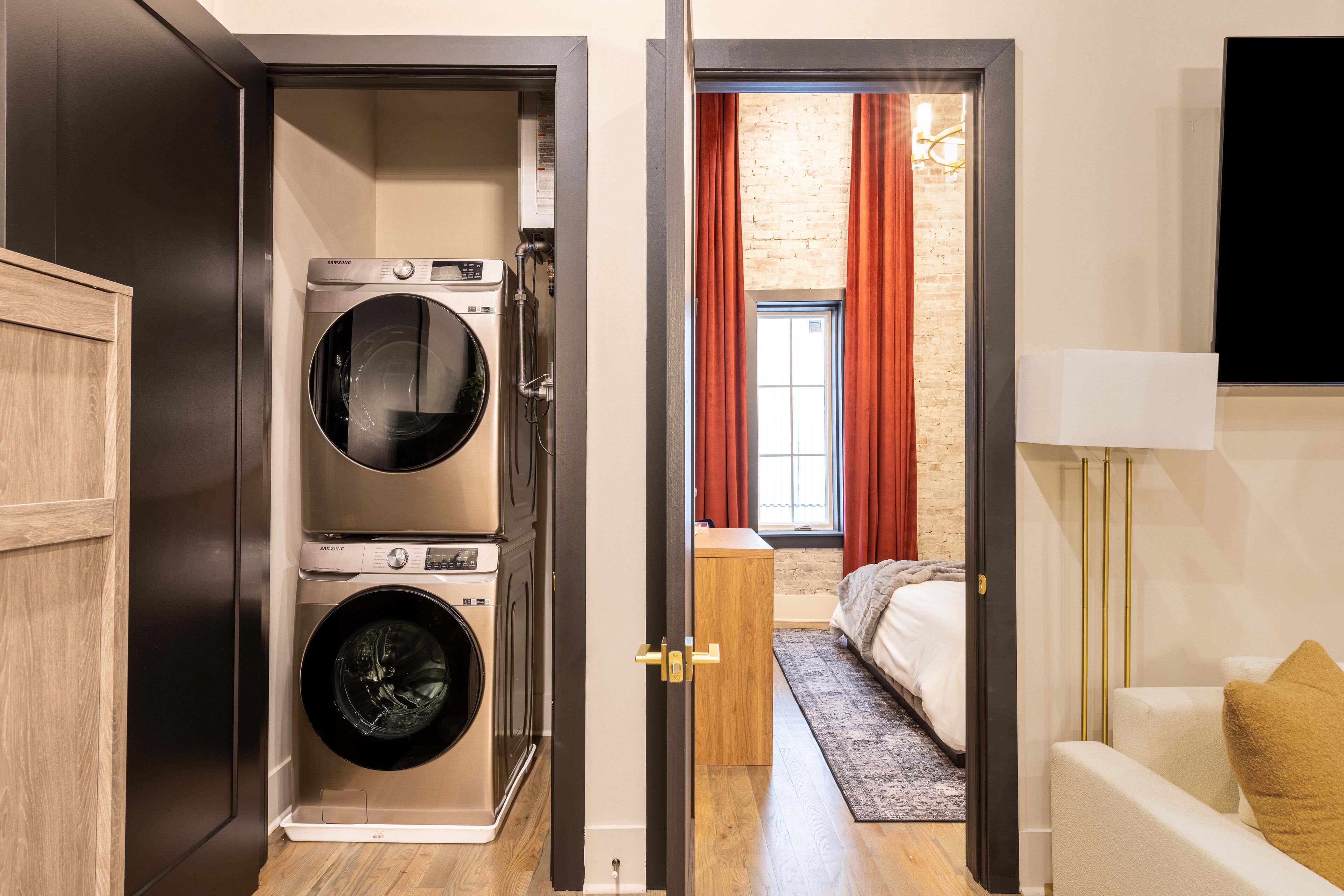 Une buanderie équipée de deux machines à laver, d’une armoire en bois et d’une porte donnant sur une chambre à coucher avec un lit, une table de chevet et une lampe.