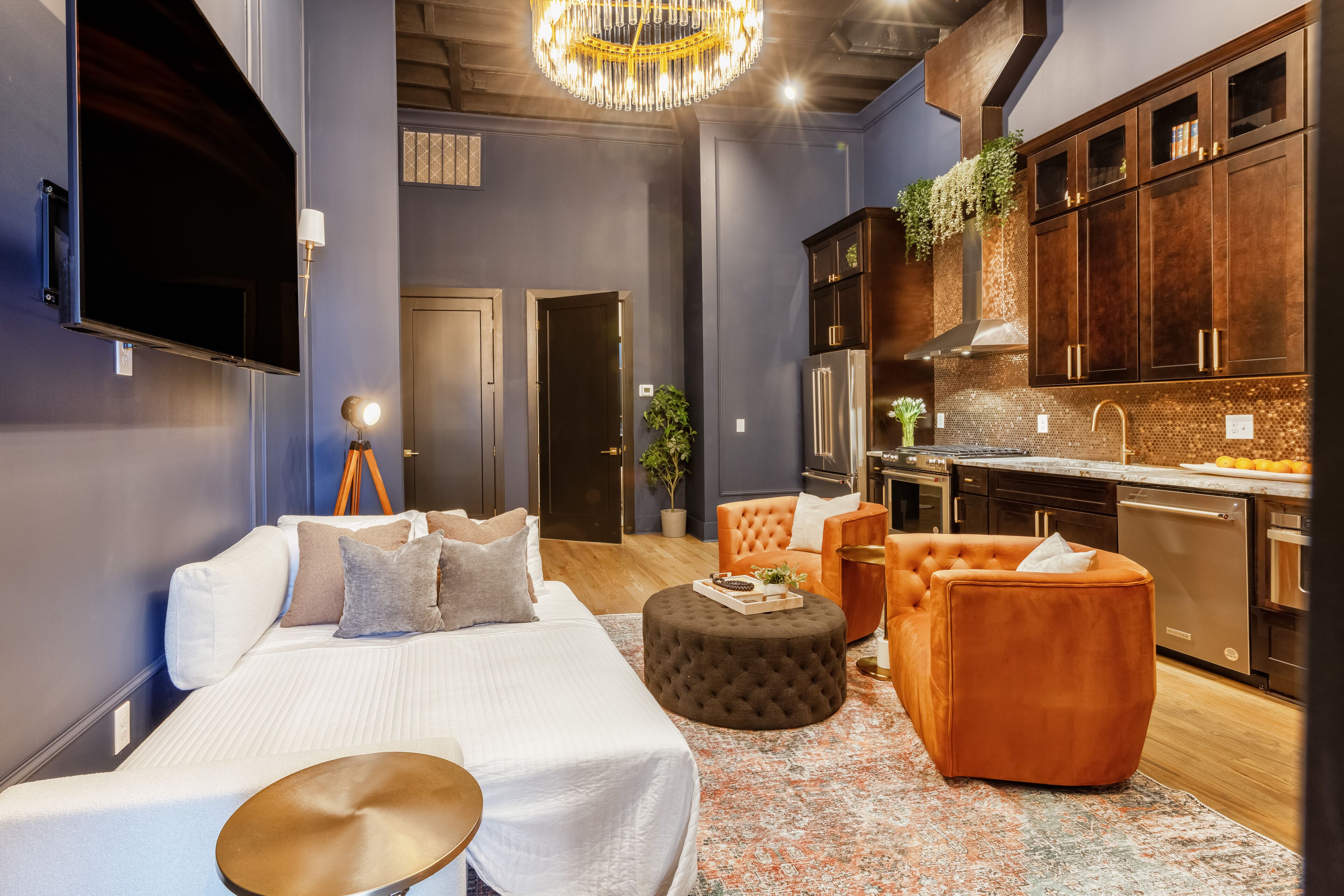 Een moderne slaapkamer met een bed, een voetenbankje en een zithoek, een keuken met donker houten kastjes en roestvrijstalen apparatuur, en een grote kroonluchter.