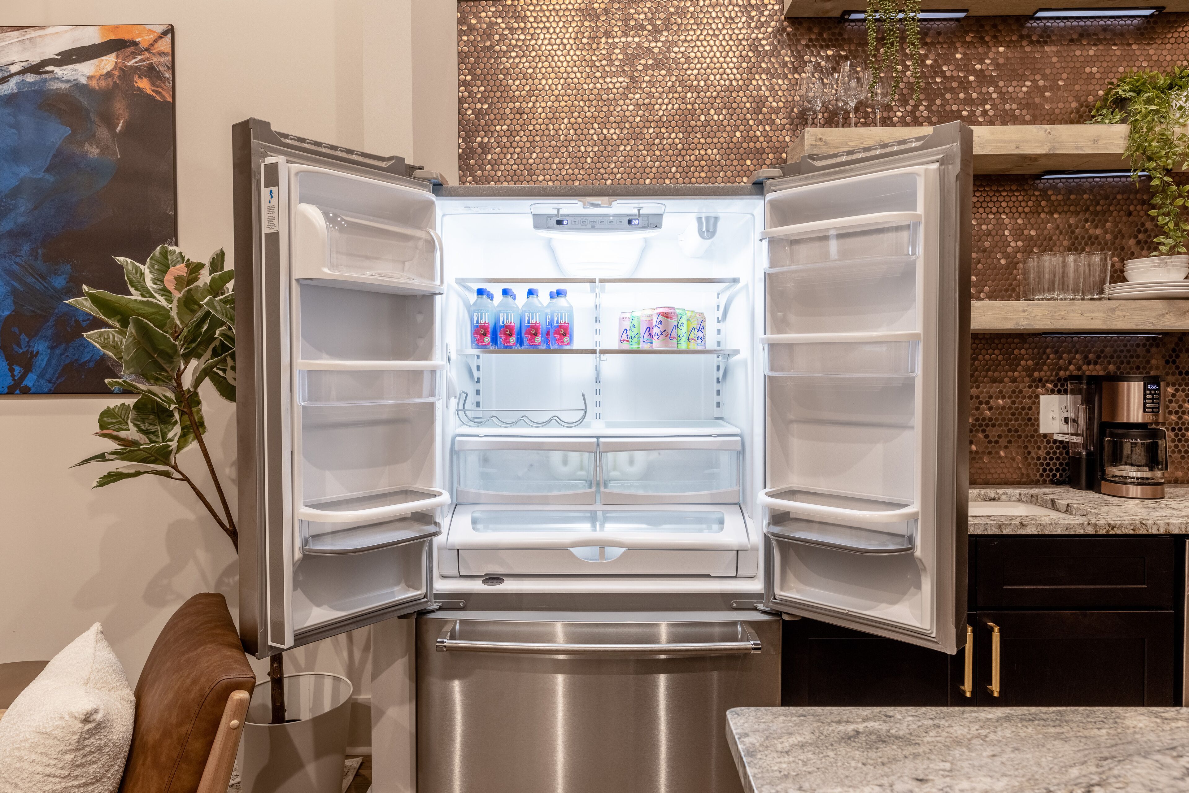 Geladeira aberta com garrafas de água e lanches, uma planta e uma bancada de cozinha com uma cafeteira.