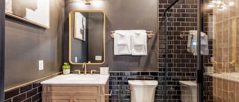 Een moderne badkamer met een donkere betegelde muur, een houten wastafelmeubel, een wit toilet en een douchecabine met glazen wanden.
