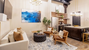 A modern living room with a sofa, ottoman, and chairs, a kitchen with dark cabinetry and stainless steel appliances, and a large abstract painting on the wall. - Union Street Lofts (Nashville)