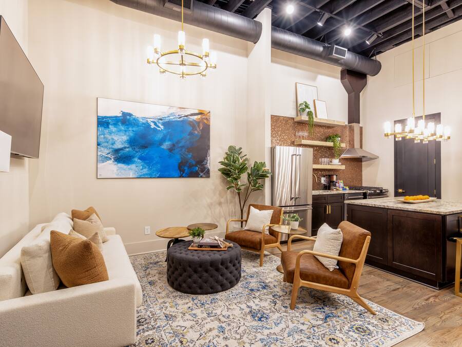 A modern living room with a sofa, ottoman, and chairs, a kitchen with dark cabinetry and stainless steel appliances, and a large abstract painting on the wall.