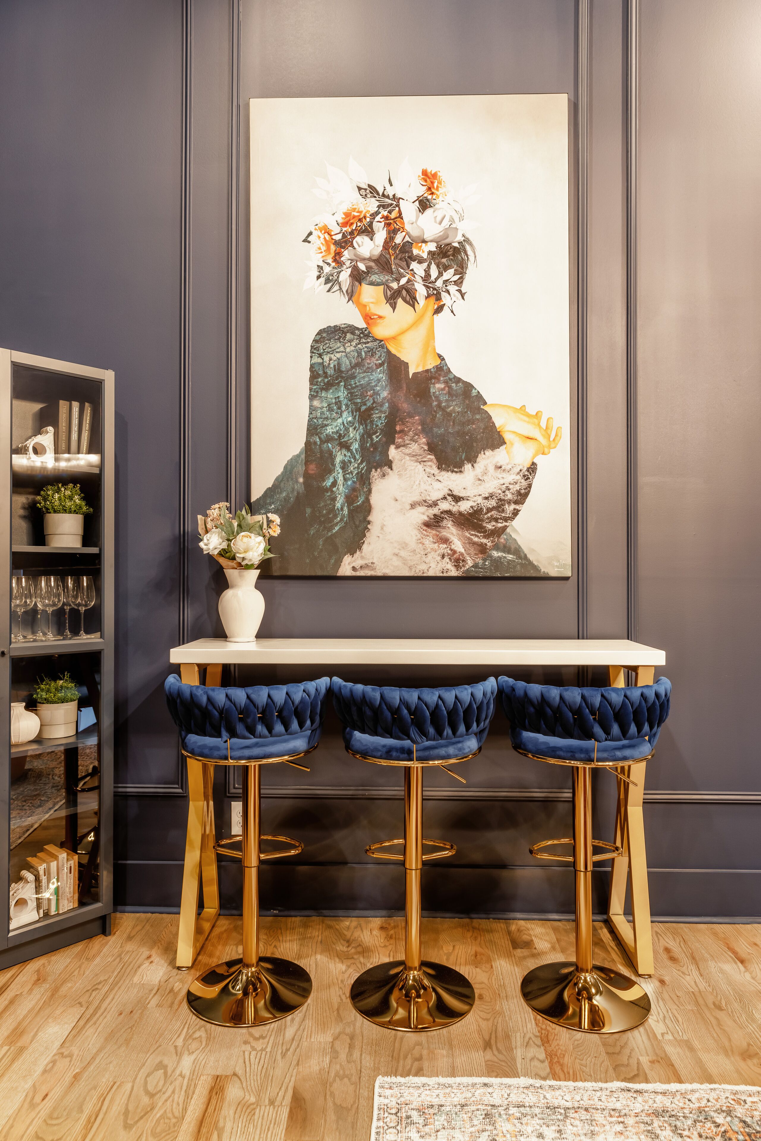 Een modern interieur met een witte tafel, blauwe fluweel-barstoelen, een foto in gouden lijst en een glazen kast met decoratieve objecten.