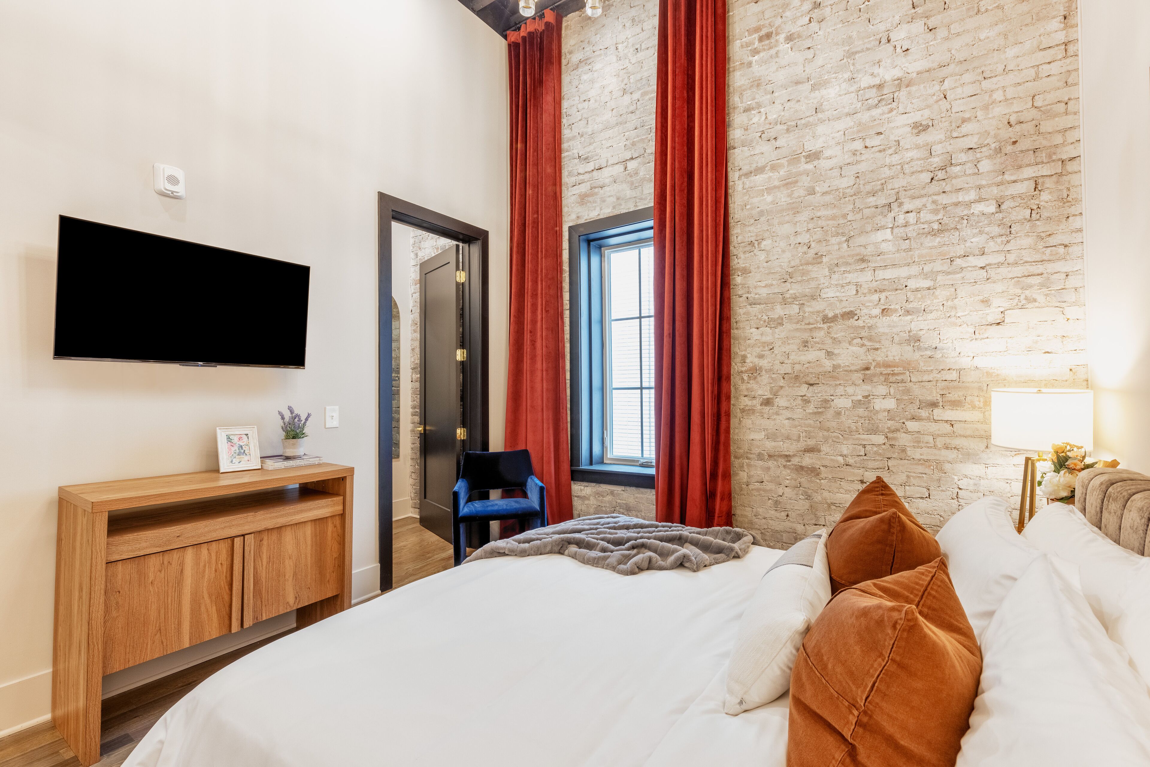 A modern hotel room with a brick wall, a flat-screen TV, a wooden console, a bed with white and brown bedding, and a window with red curtains.
