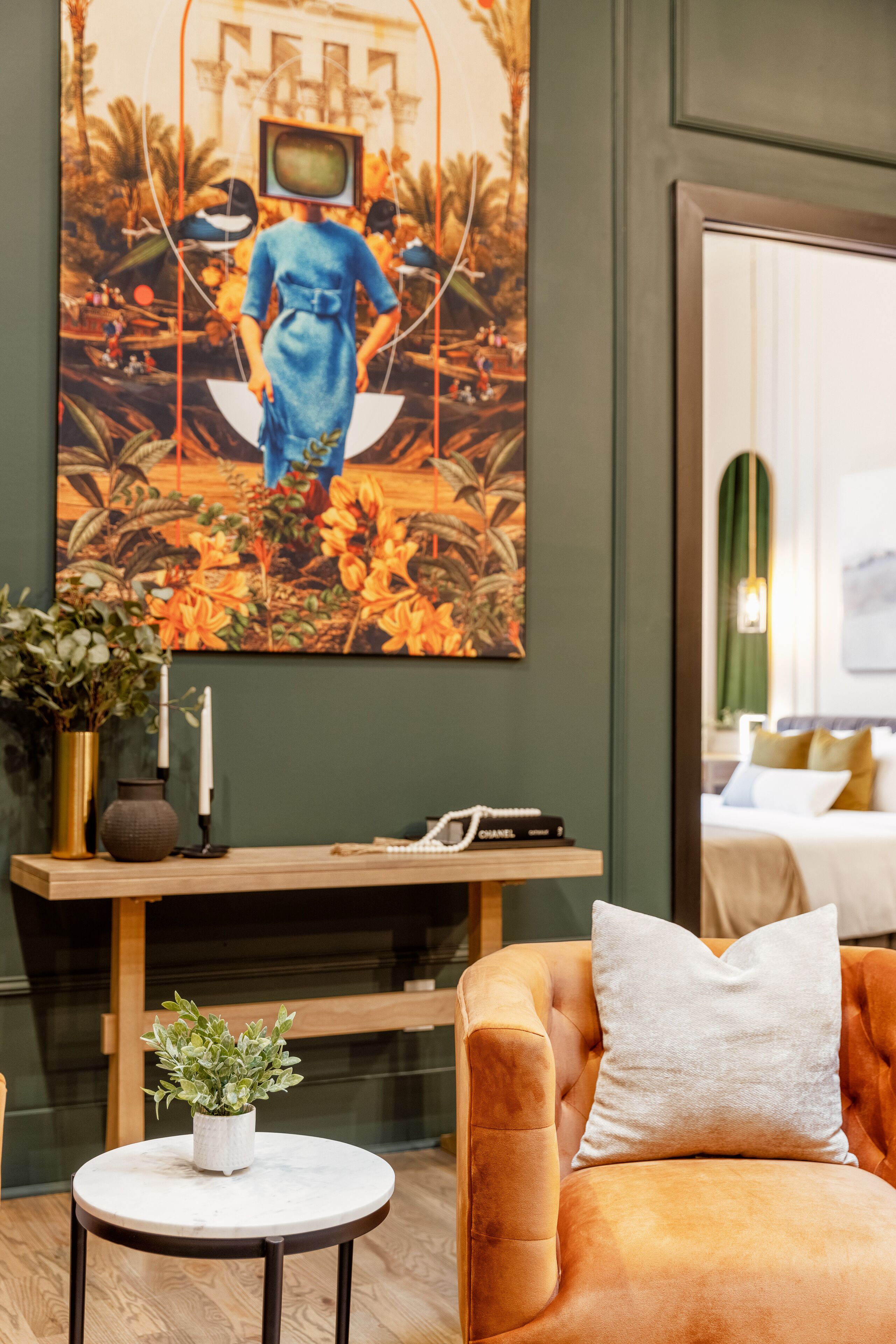 A room with a large painting of a woman on a swing, a wooden table with a plant, a cushioned armchair, and a small round table.