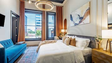 Una habitación de hotel moderna con una cama grande, un sofá azul, vistas de la ciudad y un cuadro abstracto de gran tamaño en la pared.