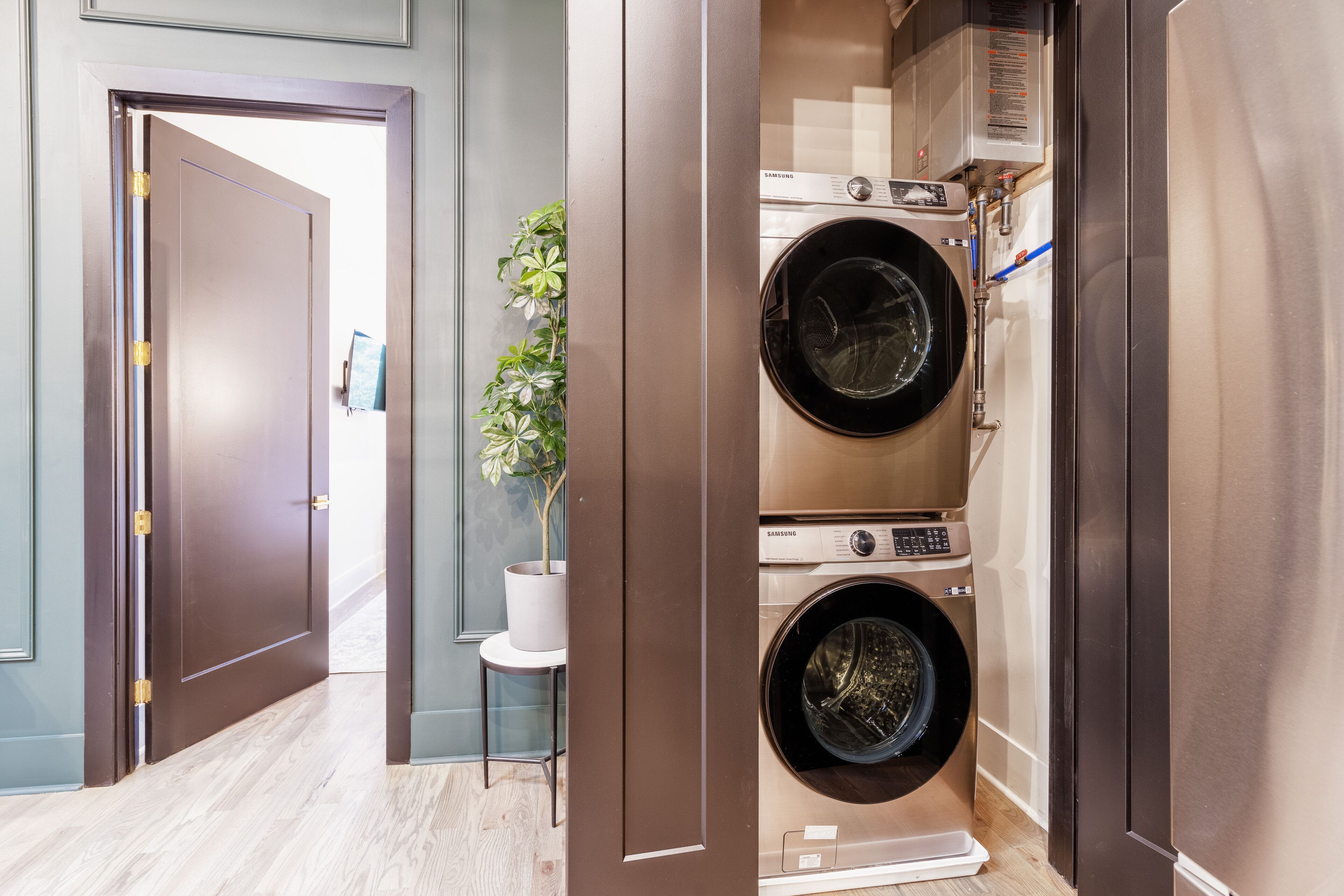 Uma área de lavanderia compacta com máquina de lavar e secadora Samsung, uma pequena planta sobre uma mesinha e uma porta que dá para outro cômodo.