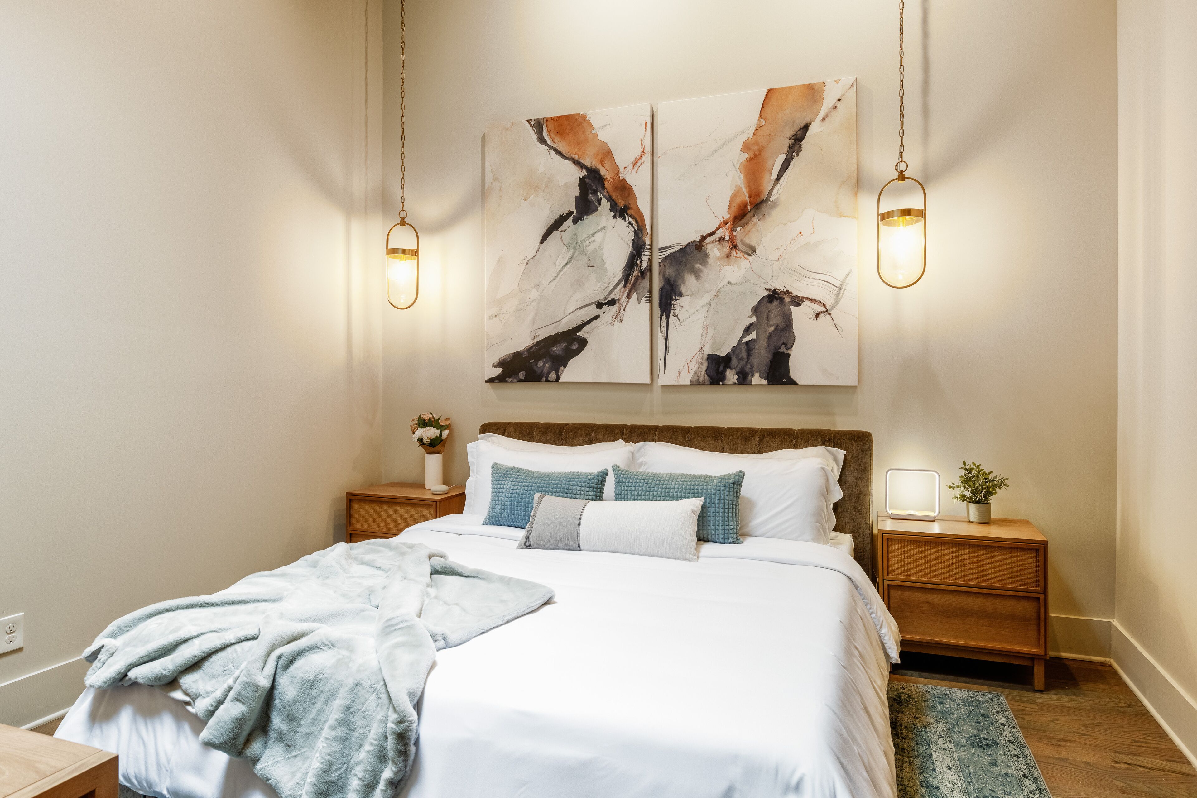 A neatly made bed with white linens and blue pillows, a bedside table with a lamp, and a wall-mounted abstract painting.