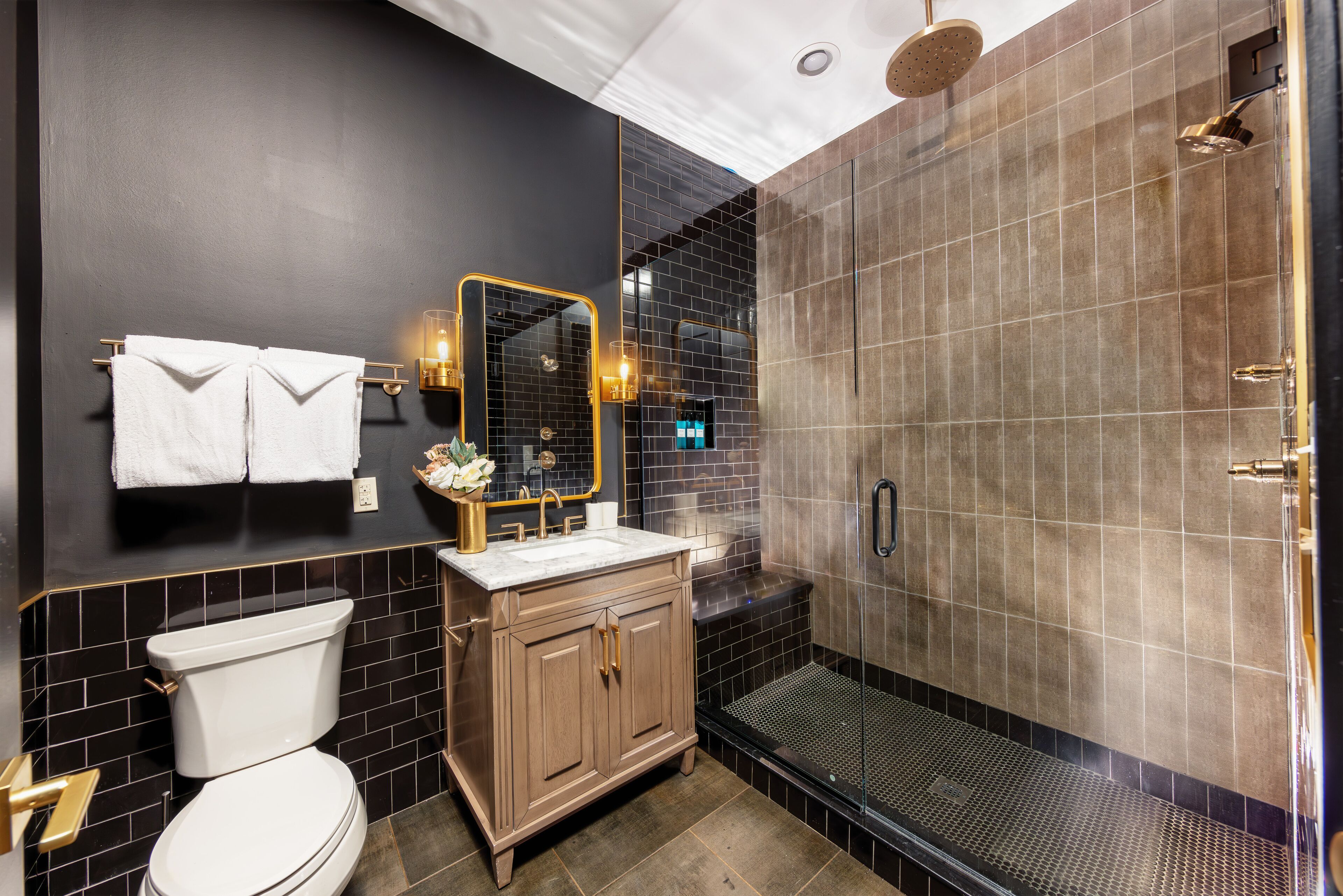 Un baño moderno con pared de azulejos negros, inodoro blanco, tocador de madera y zona de ducha con mampara de vidrio.