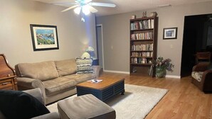 A living room with a sofa, ottoman, coffee table, bookshelf, and a ceiling fan. - Home Away From Home (St. Augustine)
