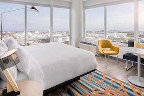 A modern hotel room with a large bed, a desk, a chair, and a view of the cityscape.