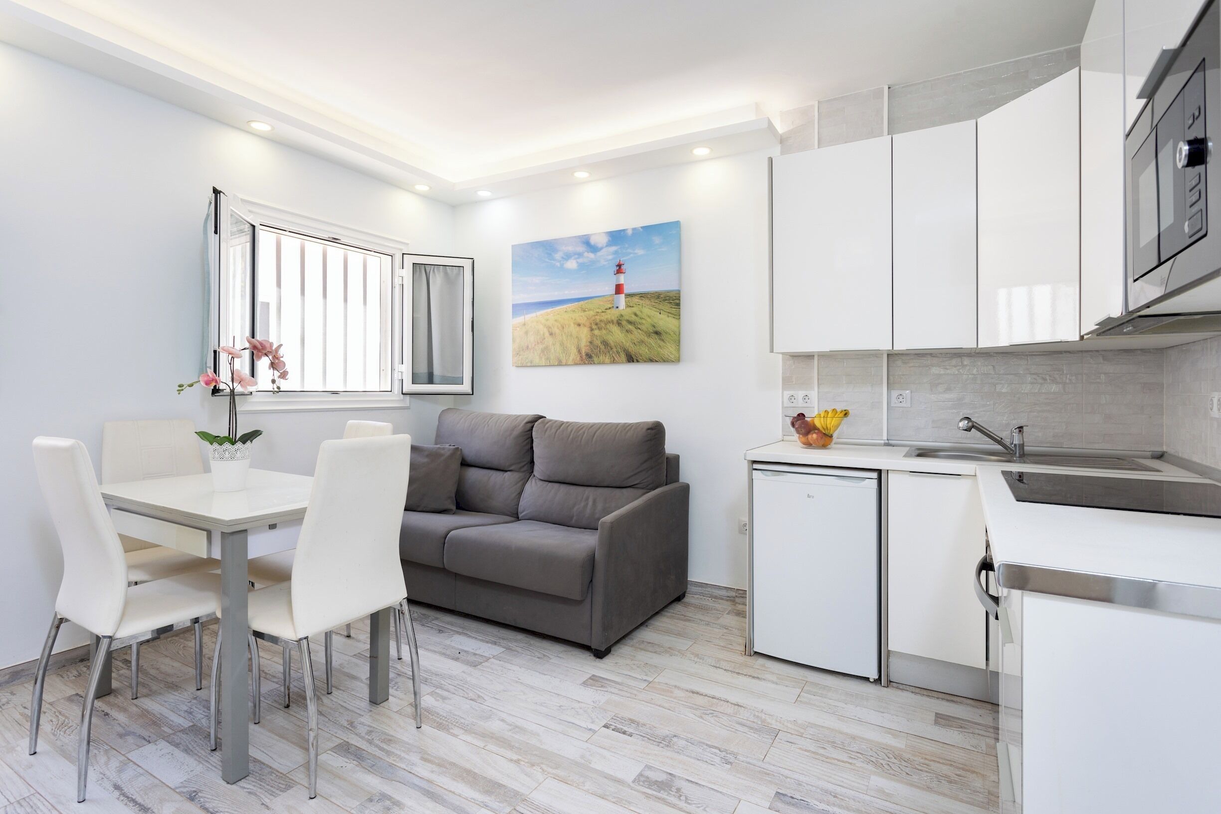 Une cuisine moderne avec des meubles blancs, un canapé gris, une table à manger avec des chaises et un petit réfrigérateur.