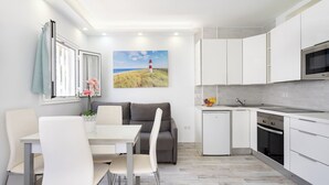 A modern kitchen with white cabinets, a built-in oven, and a microwave. There is a dining area with a table and chairs, and a sofa. A painting of a lighthouse is on the wall. - Home2Book Chill & Sun Colina Blanca & Pool, Adeje (Adeje)