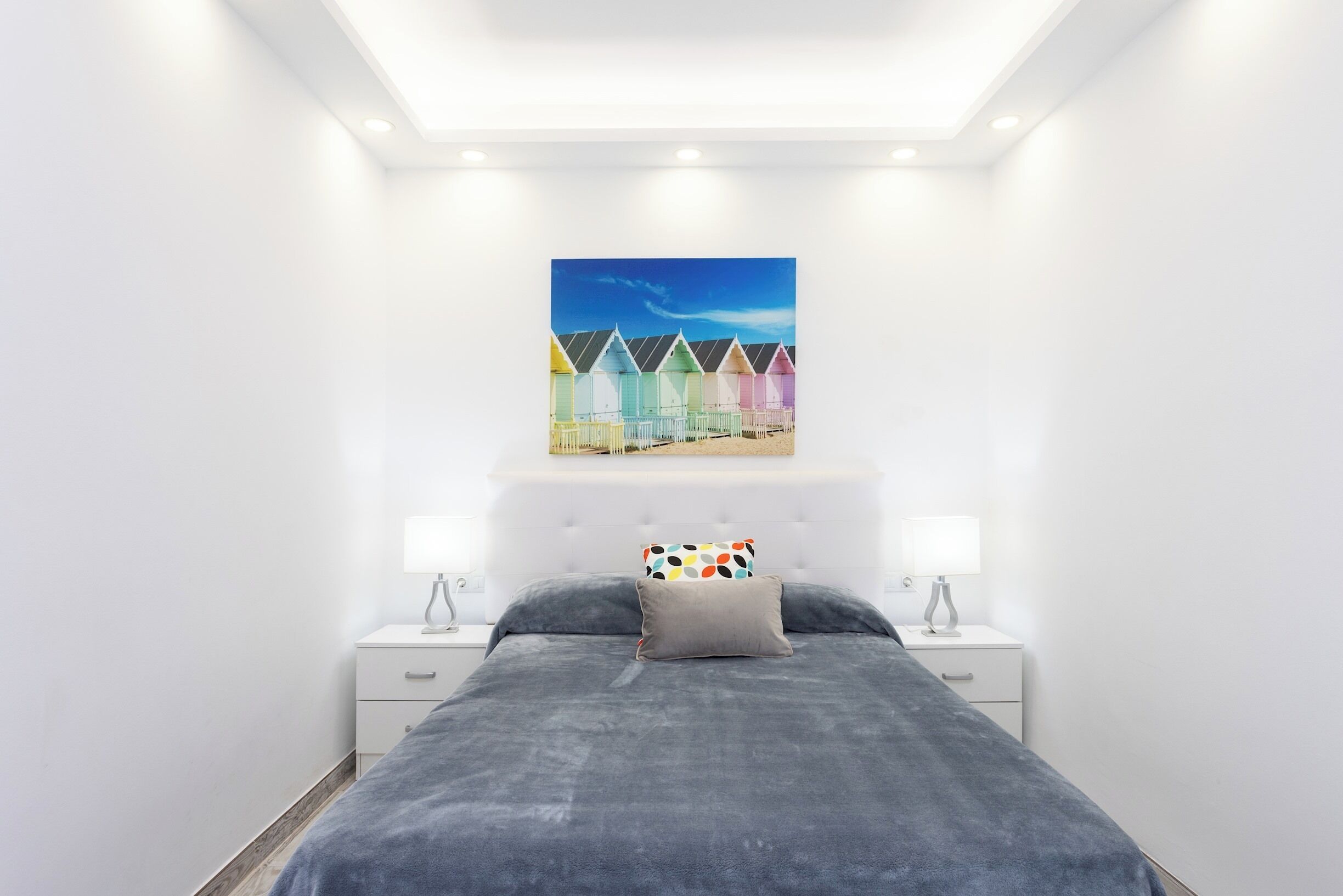 A bedroom with a bed, two bedside tables, a nightstand, a ceiling light, and a wall-mounted artwork of colorful beach huts.