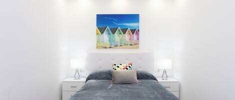 A bedroom with a bed, two bedside tables, a nightstand, a ceiling light, and a wall-mounted artwork of colorful beach huts.