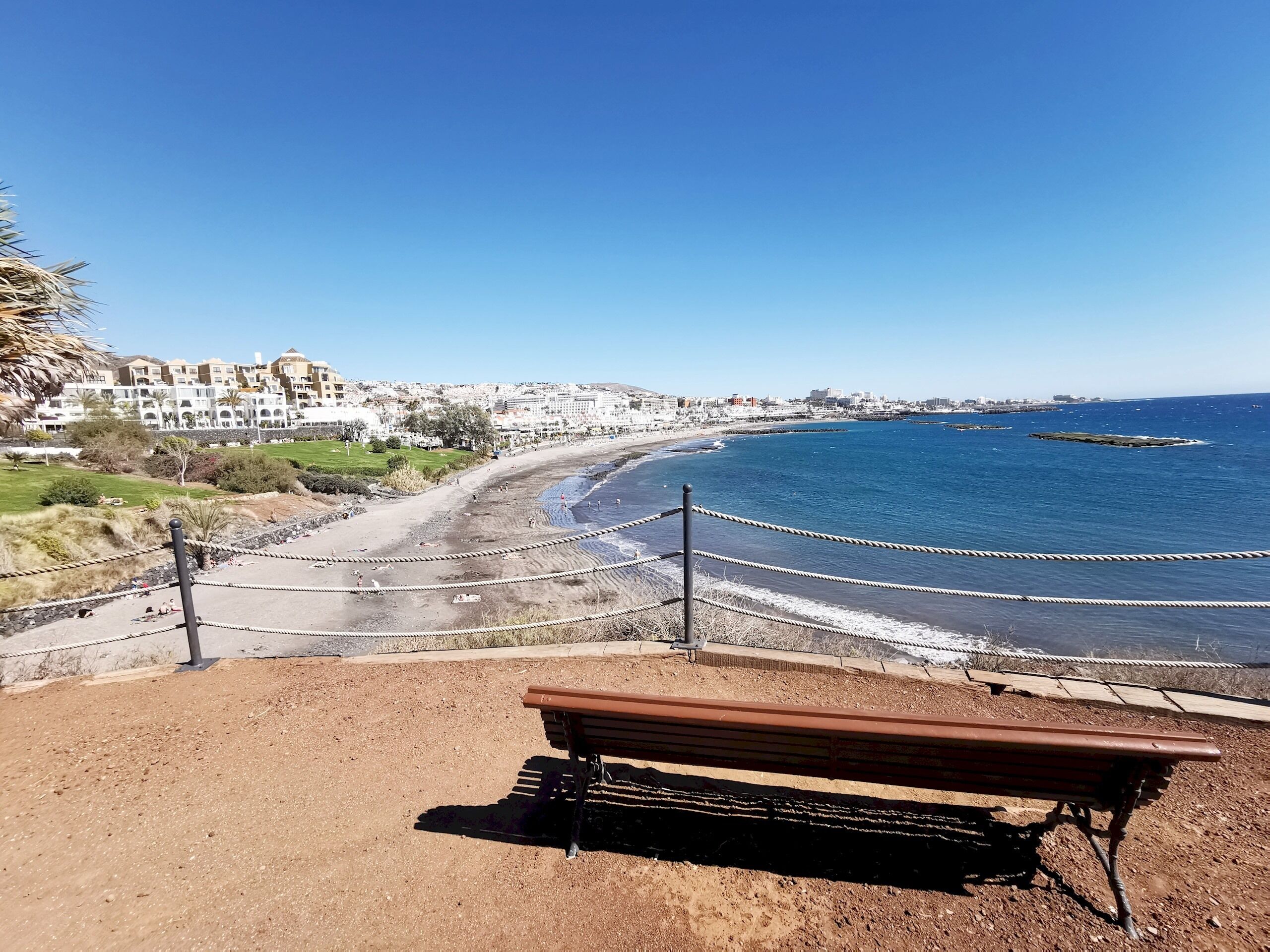 A coastal view with a sandy beach, a wooden bench, and a rope barrier.