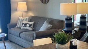 Una sala de estar acogedora con un sofá gris, una mesa de centro y una mesa de comedor. Hay dos lámparas en la mesa, una planta en maceta y una pared con anclas decorativas.