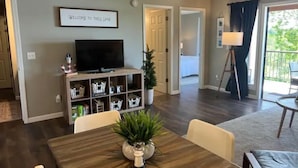 Una sala de estar con televisor, una estantería, una planta en maceta y un comedor con mesa y sillas.