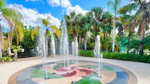 Una fontana circolare con getti d'acqua multipli, circondata da un pavimento colorato in mosaico, situata in un parco con palme e cielo sereno.