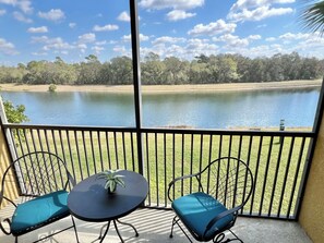 Un balcon avec deux chaises et une table, donnant sur un lac paisible et une zone verdoyante parsemée d’arbres.