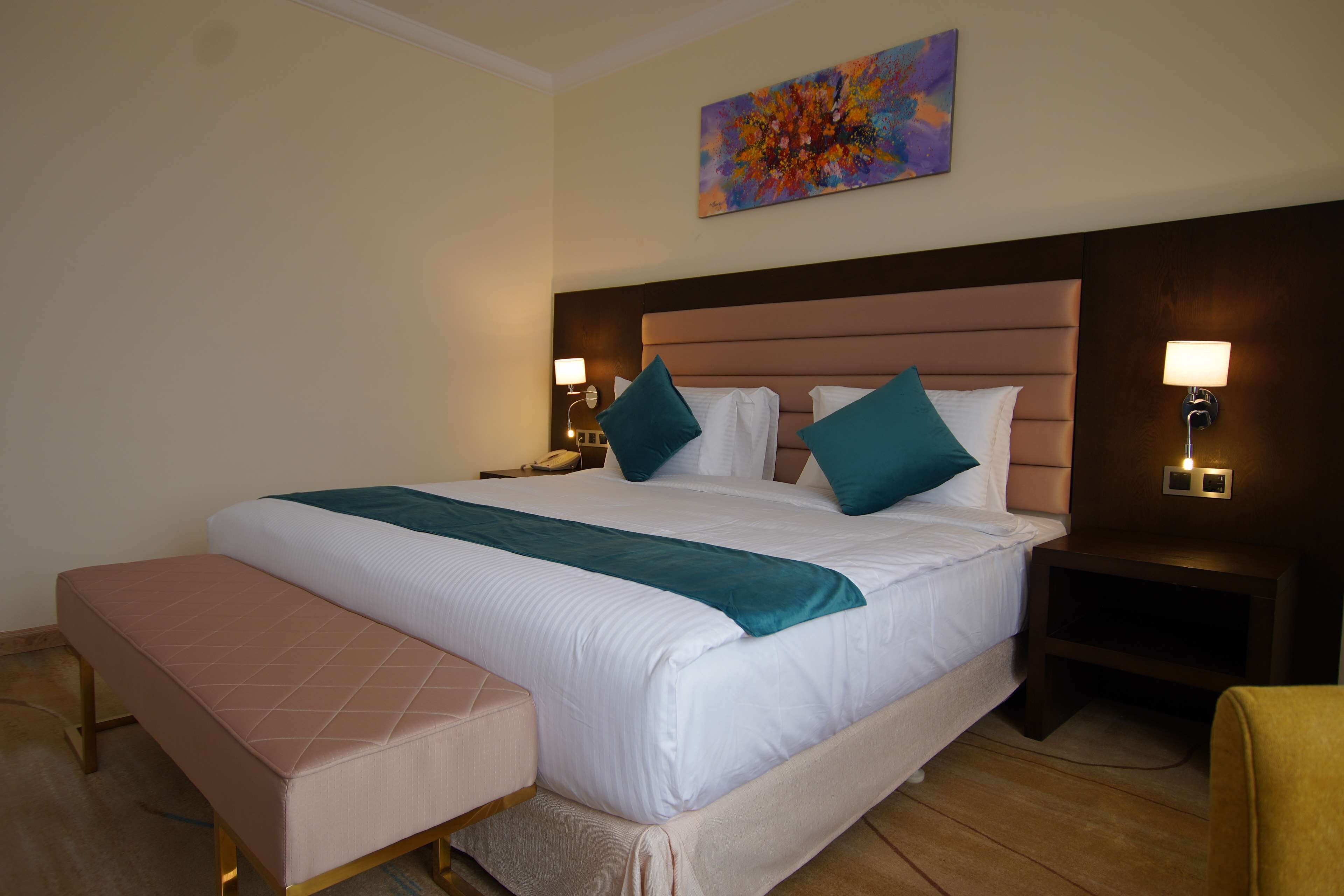 Une chambre d’hôtel avec un grand lit, deux tables de chevet avec des lampes, un banc et un tableau abstrait aux couleurs vives accroché au mur.