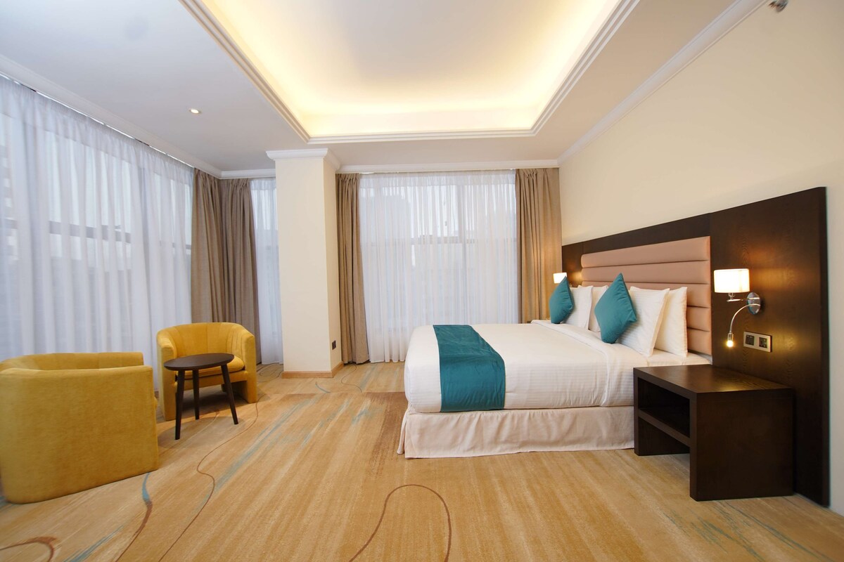 A hotel room with a large bed, a bedside table with a lamp, a small round table, a yellow armchair, and a large window with curtains.