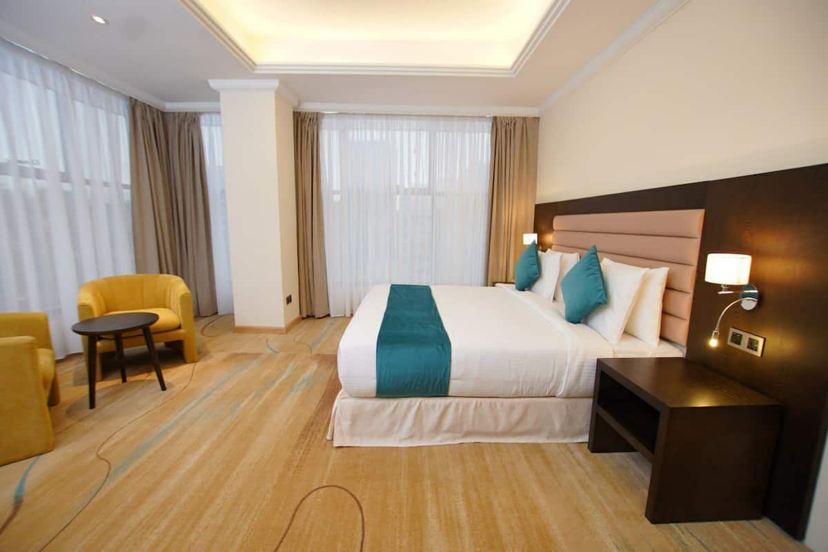 A hotel room with a large bed, a bedside table with a lamp, a small round table, and a yellow armchair.
