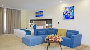 Habitación de hotel con una cama, un sofá azul, una mesa de centro con flores y obras de arte en las paredes.