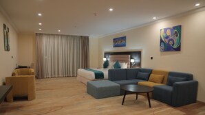 Una sala moderna con un sofá modular, un reposapiés y una pequeña mesa redonda. Se observa una cama con cabecera y un ventanal grande con cortinas.