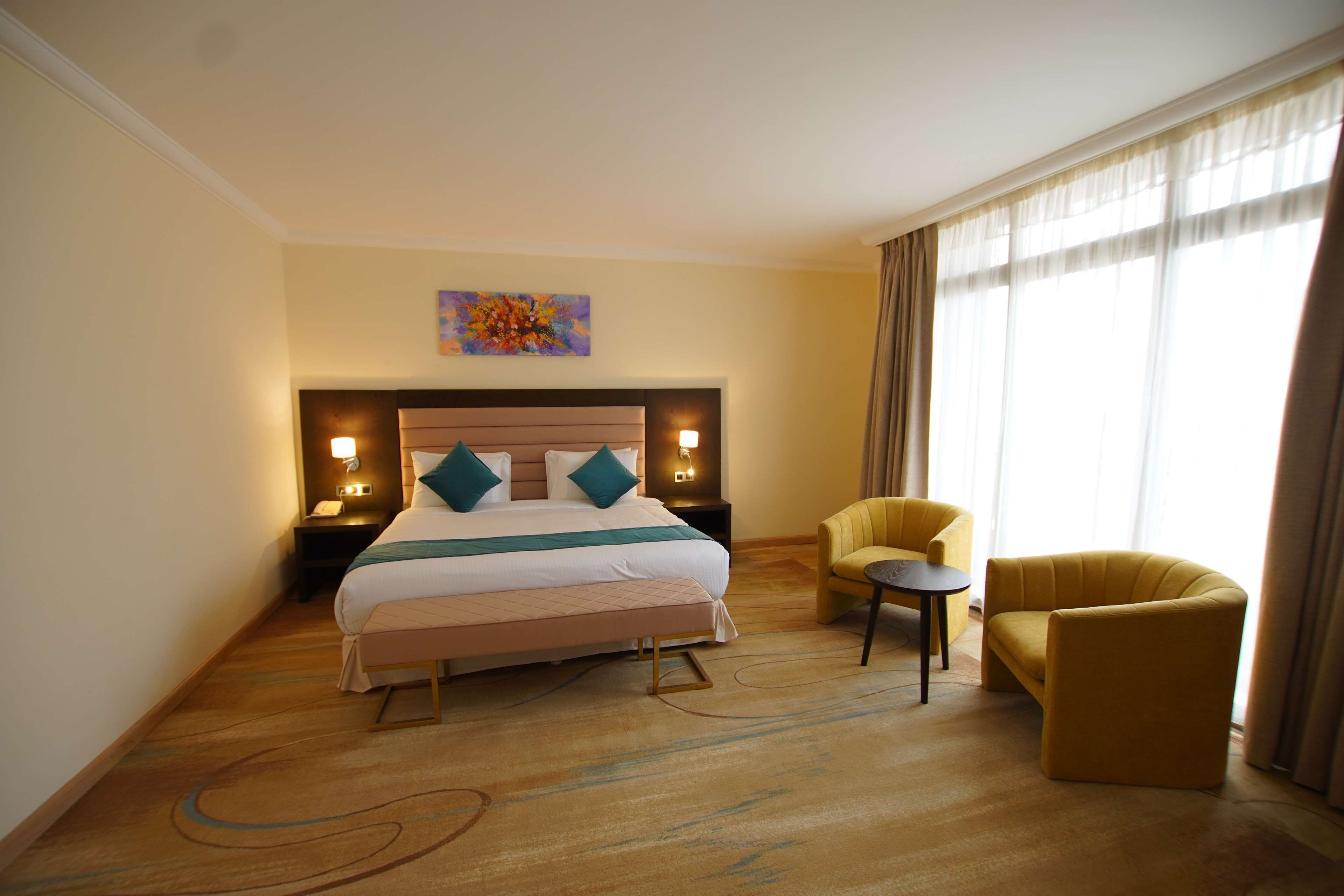 A hotel room with a large bed, two bedside tables with lamps, a small round table, two yellow armchairs, and a large window with curtains.