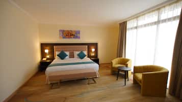 A hotel room with a large bed, two bedside tables with lamps, a small round table, two yellow armchairs, and a large window with curtains.