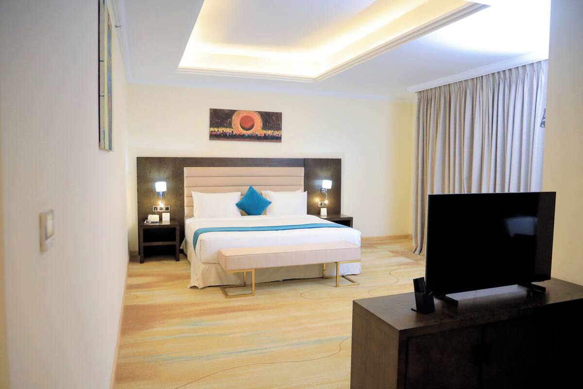 A hotel room with a bed, bedside lamps, a flat-screen TV, and a painting on the wall.