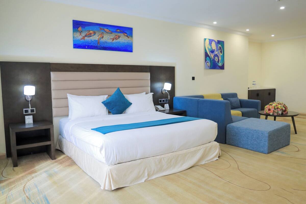 A hotel room with a large bed, a blue accent pillow, a blue sofa, a small table with a vase, and artwork on the walls.