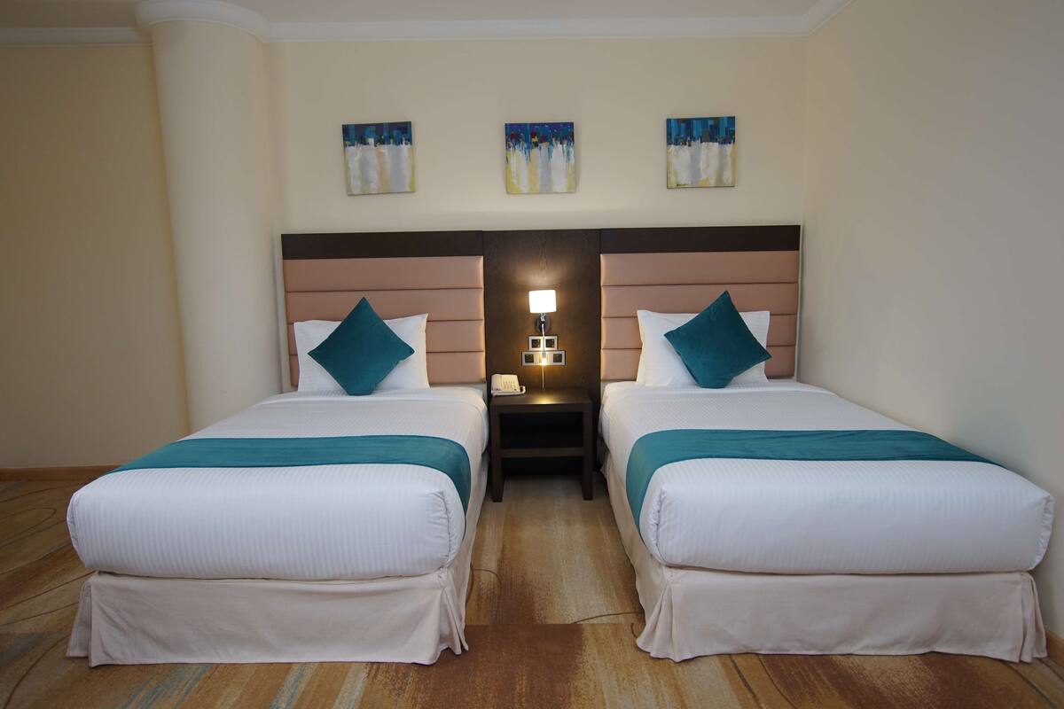 A hotel room with two single beds, a nightstand with a lamp, and three wall-mounted paintings.