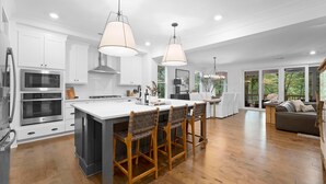 Una cocina moderna con armarios blancos, una isla central con taburetes y un comedor con vistas al exterior.