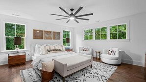 Un dormitorio moderno con una cama grande, dos sillones blancos, un ventilador de techo y ventanas amplias con vistas a la vegetación.