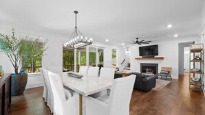 Una sala de estar moderna con chimenea, un comedor con mesa y sillas blancas, y un ventanal con vistas a zonas verdes.