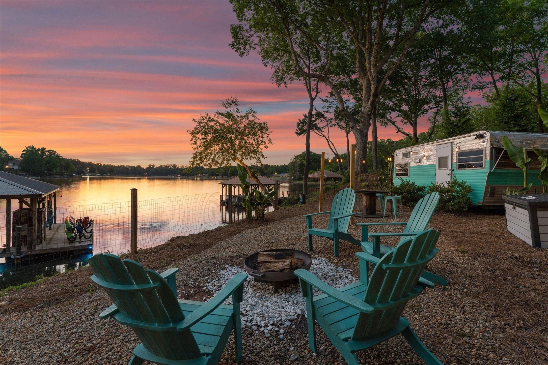 Un entorno junto al lago con un brasero, sillas Adirondack y una caravana antigua.