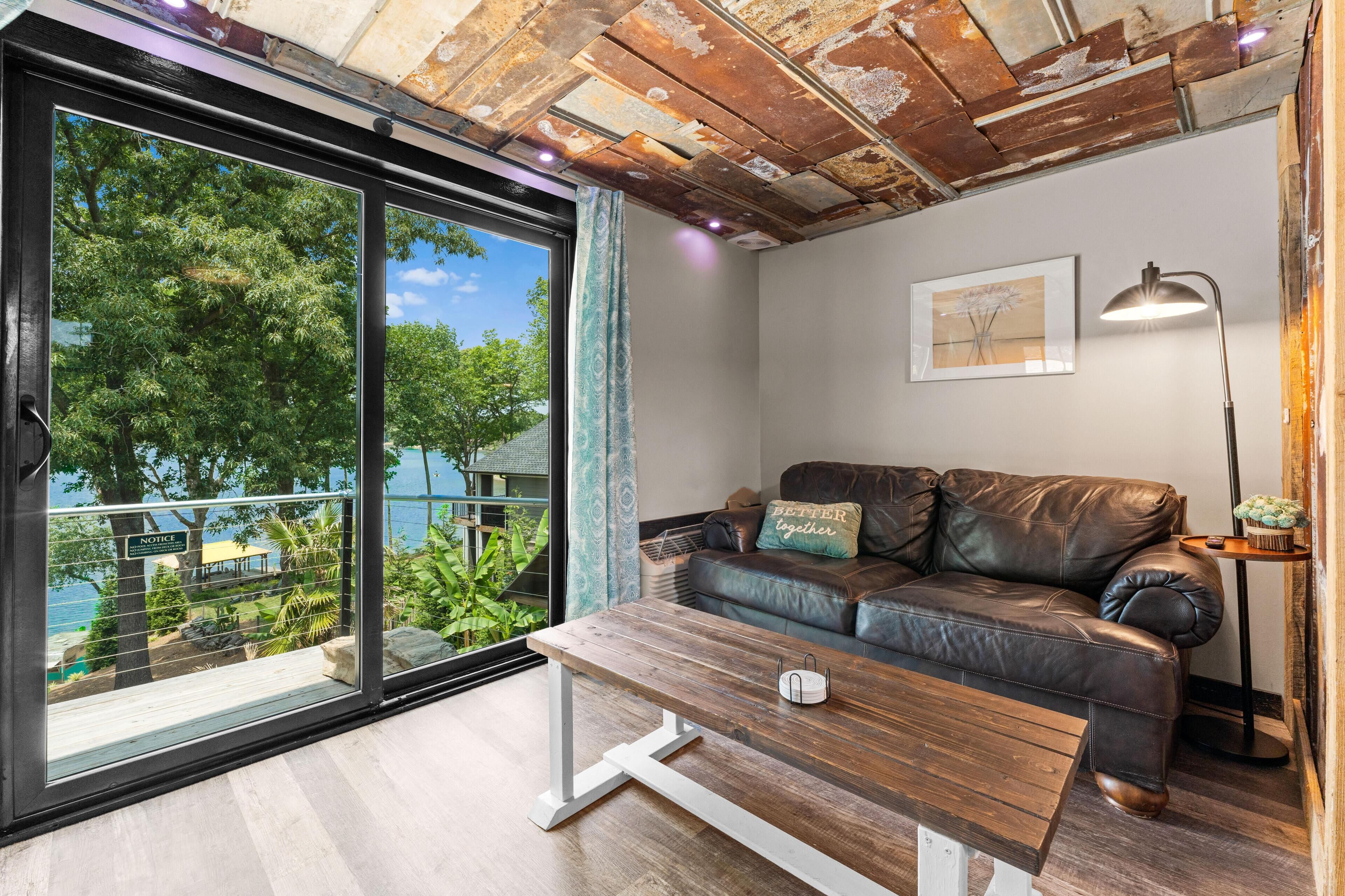 A modern living room with a leather sofa, a wooden coffee table, and a view of a balcony with greenery and a body of water.