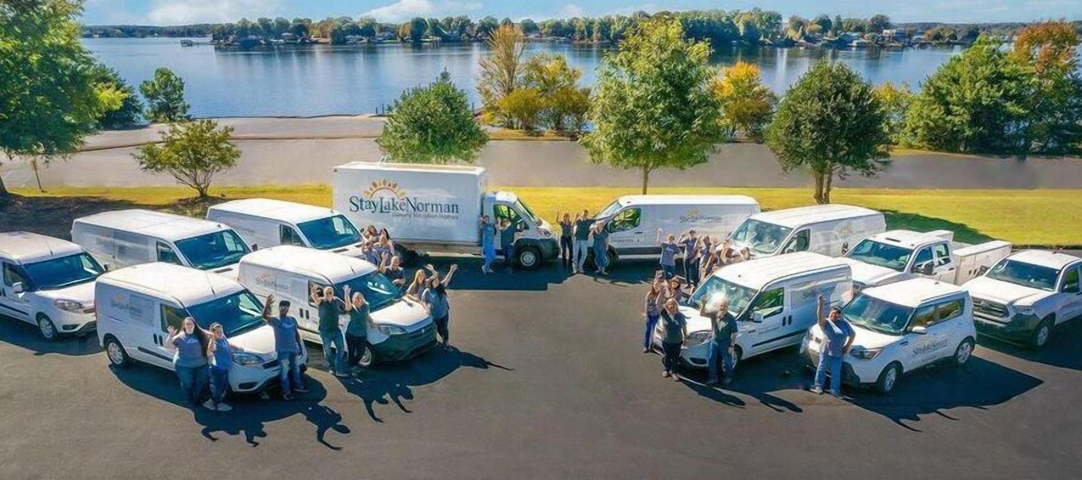 Un grupo de vehículos recreativos estacionados en un estacionamiento, con un lago y árboles al fondo.
