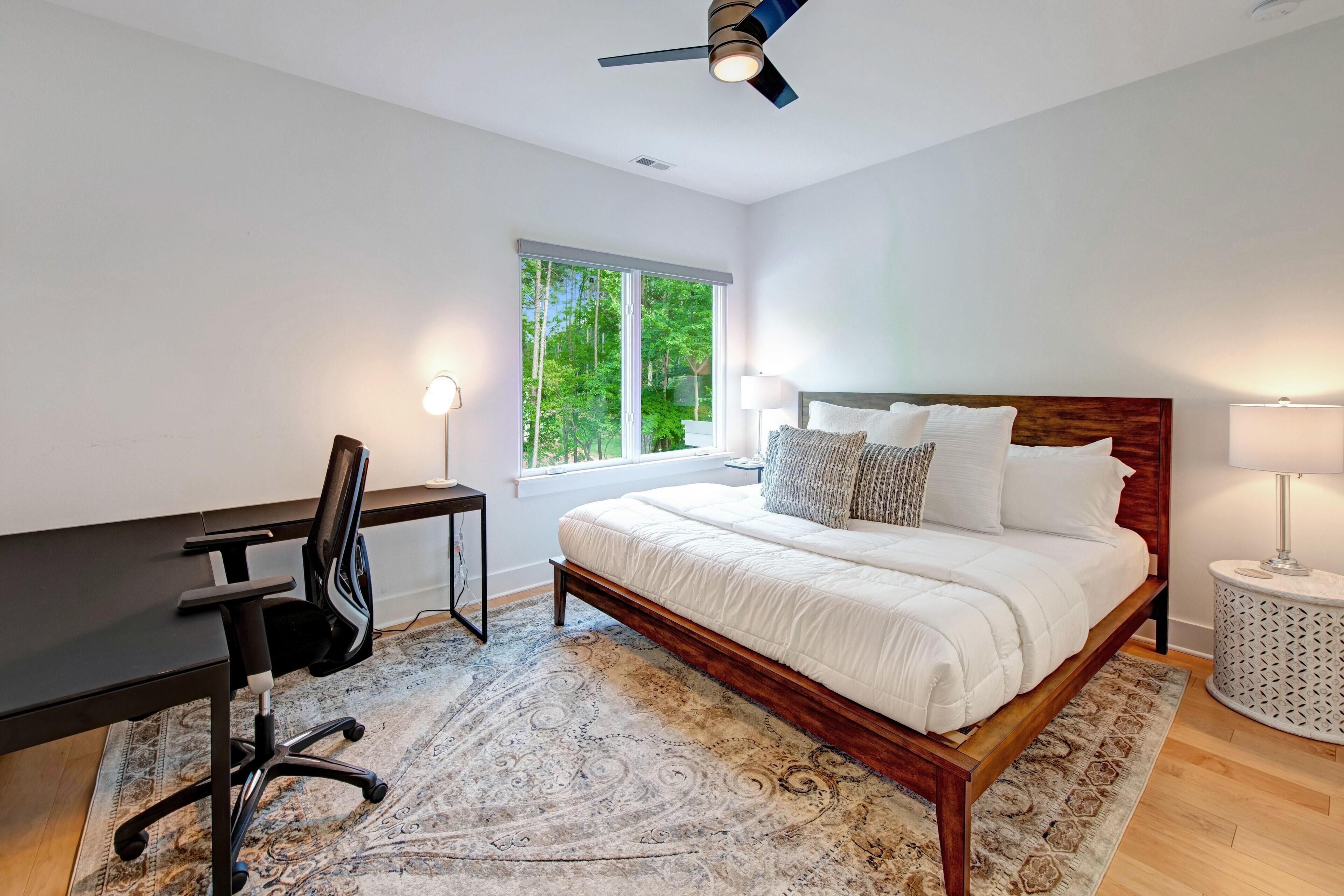 A bedroom with a bed, desk, chair, and a window with a view of greenery.
