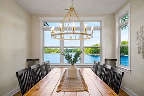 Une salle à manger avec une table en bois, des chaises noires, un lustre et une vue sur un lac, visible à travers de grandes fenêtres.