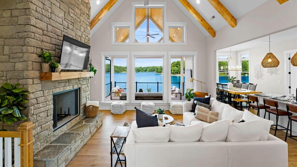A spacious living room with a stone fireplace, wooden beams, a ceiling fan, and large windows overlooking a lake. - Hogan Crest (Mooresville)
