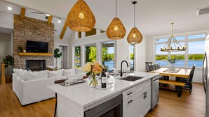 A modern kitchen with a central island, white cabinetry, and a stone fireplace. There is a dining area with a wooden table and chairs, and a large window offering a view of the outdoors. - Hogan Crest (Mooresville)