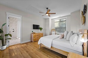 Une chambre à coucher avec un lit, un plancher en bois, un ventilateur de plafond, une fenêtre avec des stores, une lampe de chevet et un téléviseur fixé au mur.