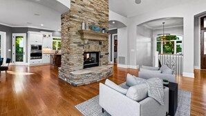 Un salón amplio con chimenea de piedra, un sofá gris y una cocina al fondo.
