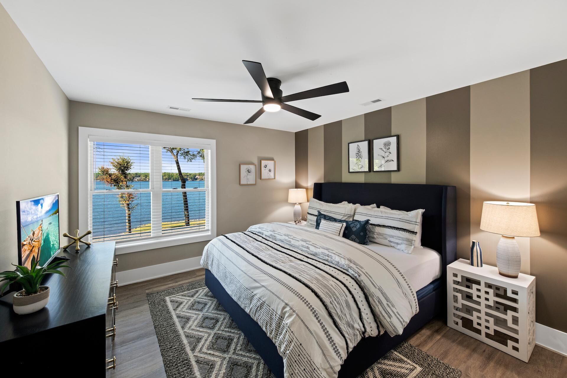 A bedroom with a bed, a ceiling fan, a window with a view of the water, and a nightstand with a lamp.