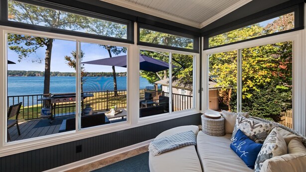 A sunroom with large windows overlooking a lake, a deck with outdoor furniture, and a cozy seating area with cushions. - Channel Marker (Mooresville)