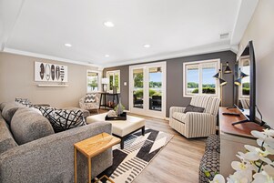 A modern living room with a grey sofa, a striped armchair, and a wooden coffee table. There are large windows with a view of greenery outside.