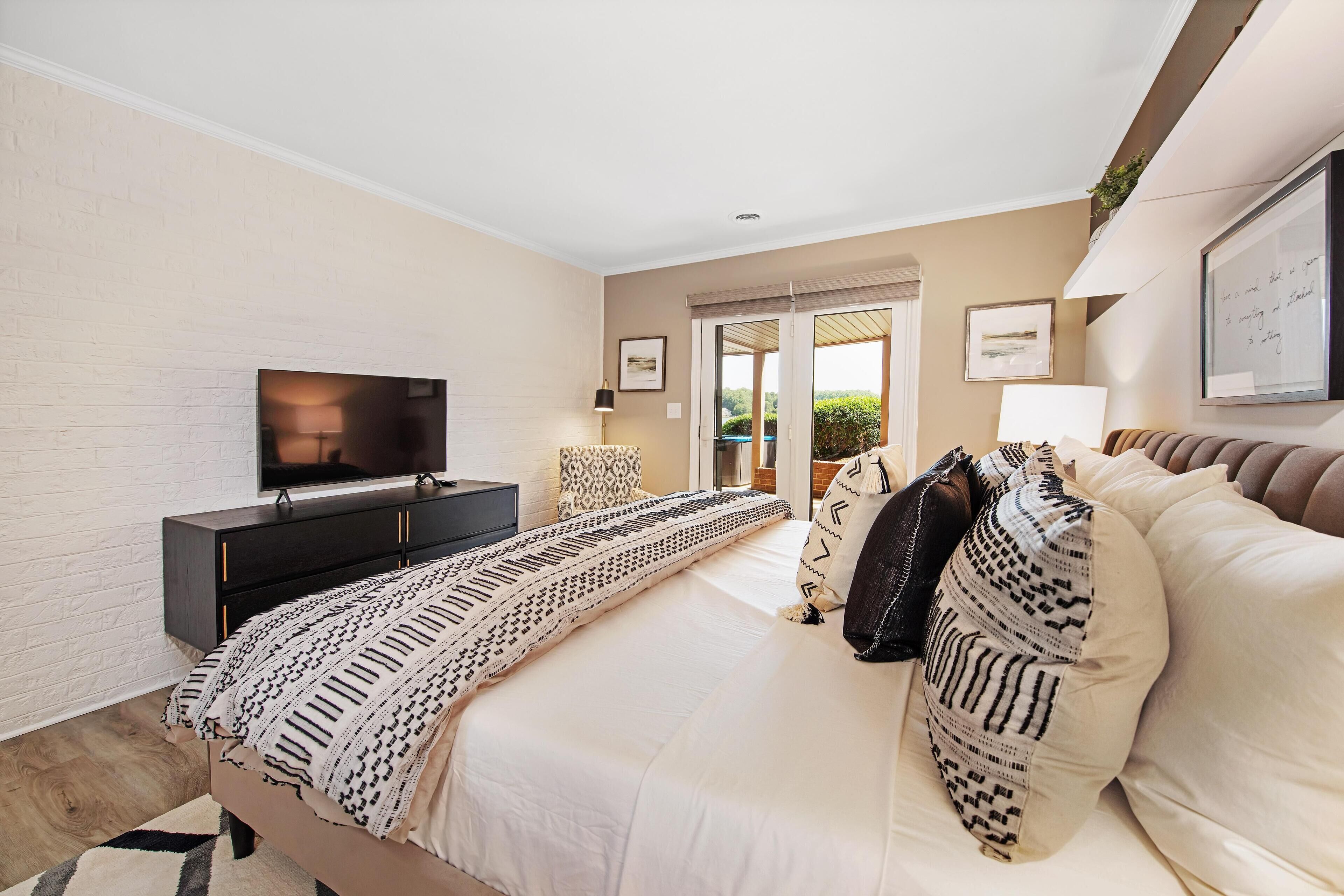 A modern bedroom with a large bed, a TV on a black cabinet, and a view of the outdoors through a glass door.